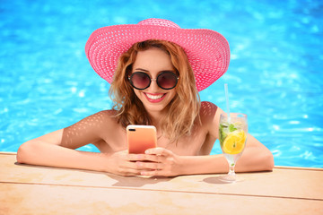 Beautiful young woman with glass of lemonade and mobile phone in swimming pool