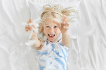 Playful freckled adorable girl with blue eyes, cathing feathers after tearing into pieces pillow, having fun before going to kindergarten. Small girl playing with feathers on white bedclothes.