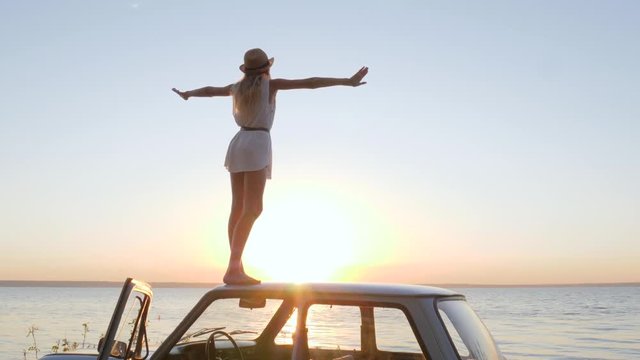 female raises arms at sunset on automobile at background sea in slow motion, happy journey, emotion happiness, woman stand on roof car, summer travel on classic vintage car, girl raises arms up on