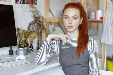 Amazing professional female artist with freckles and long ginger hair sitting at computer on white table in her modern workshop, having pensive look, deep in thoughts, absorbed with creative ideas