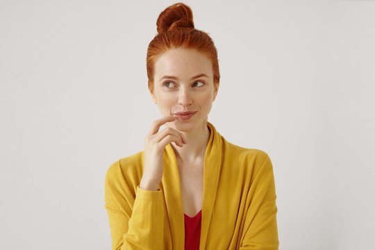 Portrait of stunning young female wearing her ginger hair in knot touching lips, looking away with sly cunning and cunning smile. Pretty girl having thoughtful dreamy look posing at studio wall