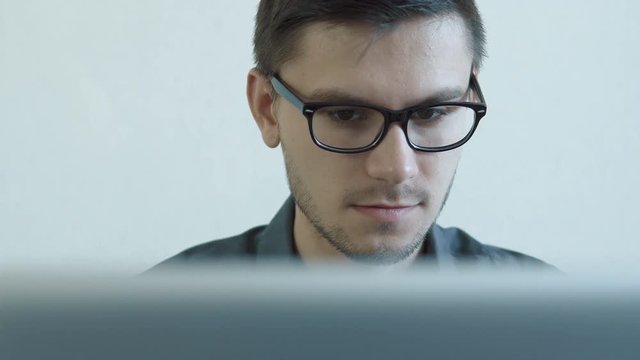 Close-up portrait of a young man wearing glasses sitting in his office in front of a monitor - working on a computer. People stock footage slider shot. 