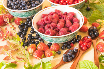 Fresh and colorful berries assortment with green leaves on the light wooden background.