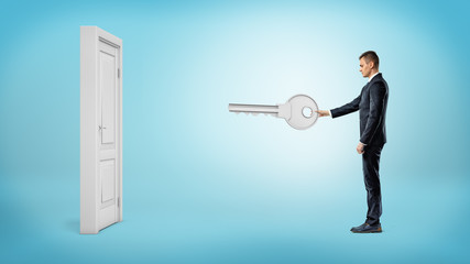 A businessman holds a giant silver key ready to open a white isolated door on blue background.
