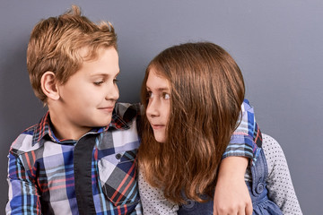 Couple of kids looking at each other. Portrait of caucasian children lovely hugging on grey background. Expression of love between little sister and brother.