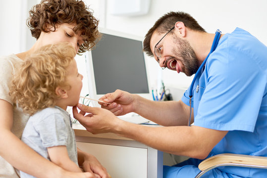 Portrait of curly child sitting on mothers lap during medical checkup, with doctor
inspecting his open mouth