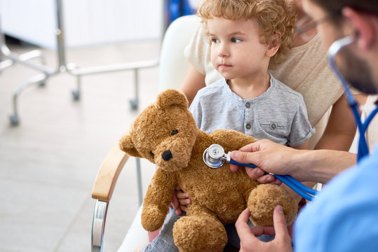 Portrait of adorable curly child in doctors office holding teddy bear toy, with pediatrician listening to heartbeat using stethoscope