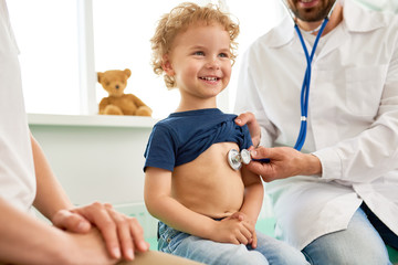 Portrait of adorable little boy visiting doctor, looking brave and smiling, holding  while pediatrician listening to heartbeat with stethoscope - obrazy, fototapety, plakaty