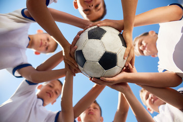Obraz premium Low angle view of boys in junior football team standing in circle holding ball together against blue sky, focus on ball