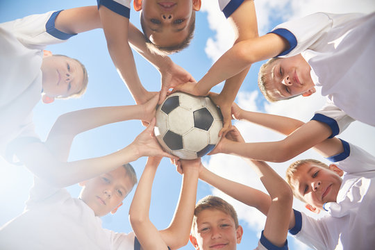 Low Angle View Of Boys In Junior Football Team Standing In Circle Holding Ball Together And Looking At Camera Against  Blue Sky