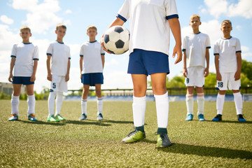 Low section portrait of football captain standing in center of field holding ball with the rest of team in background