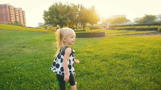 SLOW MOTION: happy girl of three years looking into the lens and running along the green grass in the city park at sunset.