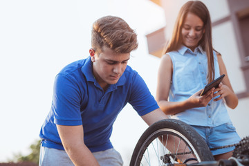 Teenager boy repair tire on bicycle , female friend standing next to him, using digital tablet for instructions, summer outdoor photo