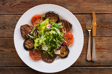 Medallions with grilled vegetables on plate. Meat, tomatoes, eggplant and green salad with grated cheese, serving in restaurant, top view