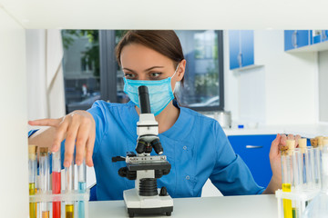 Female scientist in uniform wearing a mask is holding test tubes with liquid and making some research