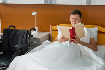 Young ill patient lying in the hospital bed and reading a book in hospital ward