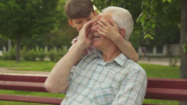 Attractive little boy shutting grandpa's eyes with his hands in the park. Handsome caucasian kid playing with his grandfather in guess who. Senior gray man sitting on the bech while his grandson