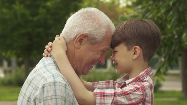 Slow motion handsome senior man and his grandson leaning their foreheads one against another. Side view of little brunette boy holding his hands behind his grandpa's neck outdoors. Attractive