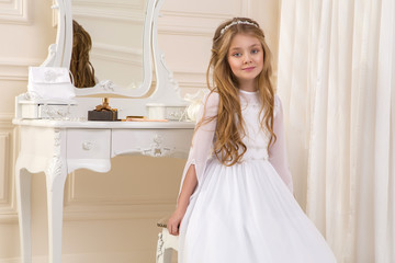 beautiful girl little princess long hair, curly and amazing blue eyes in communion dress and tiara on his head, sitting in elegant palatial room with a dressing table with mirror in the background © marcink3333
