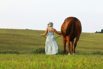 Blonde woman in summer dress walk slowly through meadow beside her horse