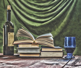 Wine in the bottle and the blue glass, books and open book on an old wooden table, closeup.
