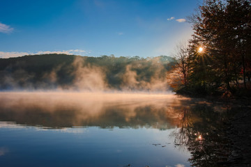 Fototapeta na wymiar sunburst at sunrise with foggy lake with autumn trees in background