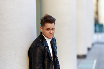 Lifestyle portrait of handsome young man with stylish haircut and smooth face wearing back leather jacket over white shirt with tie standing outdoors at column, staring at camera with serious look