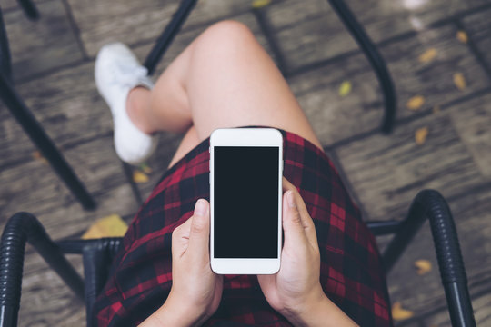 Mockup top view image of woman's hands holding white mobile phone with blank black screen on thigh with wooden floor background in vintage cafe