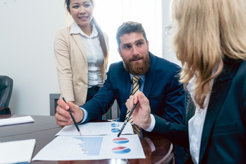Business analyst smiling while interpreting financial reports showing profit and development during meeting with his female colleagues in the office
