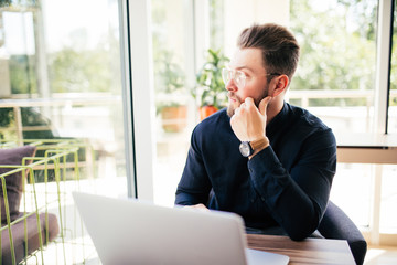 Friendly executive sitting in front of laptop in his office. Big window at the background. Looking away, daydreaming