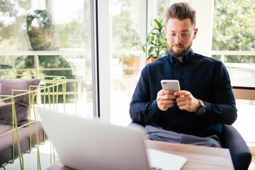 Young happy businessman smiling sitting in office with laptop while reading his smartphone. Portrait of smiling business man reading message with smartphone in office.