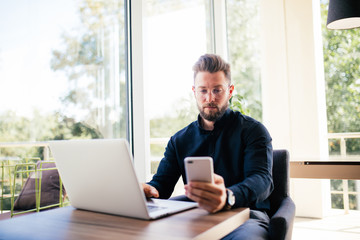 Fototapeta premium Handsome bearded man holding a mobile phone in front of laptop on wooden table with Big window at the background.