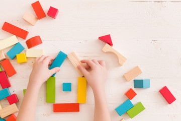 Top view on child's hands playing with colorful wooden bricks on the white table background.Kid building with geometric shapes. Learning and education concept.