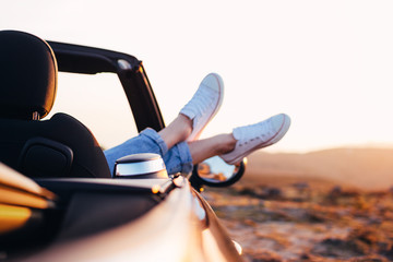 Trendy hipster woman rests and admire sunset in mountains from her cabriolet car. Happy female...