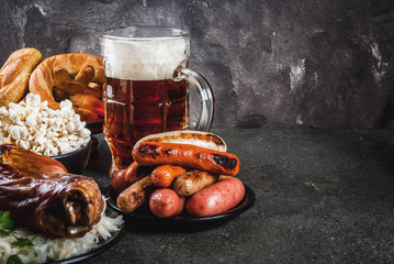 Selection of traditional German food Oktoberfest. Beer, baked pork shank, popcorn, assortment of different sausages, homemade bretzels. On a black stone background copy space
