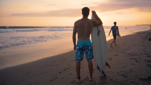 Young male surfer standing sand beach sunrise holding surfboard watching ocean waves sun lens flare. Shot taken by a handheld gimbal in 60FPS