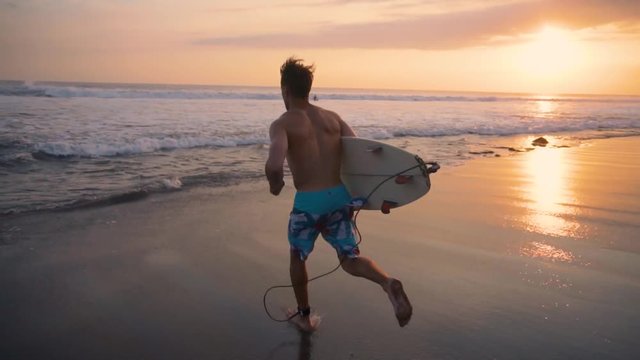 Young surfer holding board, running to go in the water at amazing golden light sunset. Happy man enjoying summer evening and seaside vacation activities. Shot taken by a handheld gimbal in 60FPS.