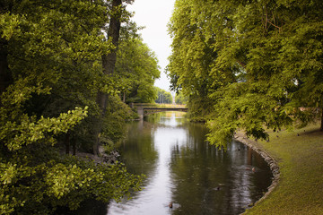 View of river and trees at Hofgarten park. Lush green space with meadows & formal flowerbeds, plus a pond, bandstand.