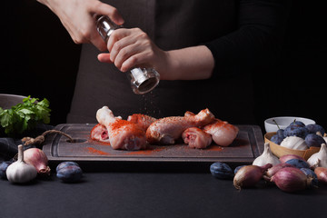 A female chef sprinkles fresh raw chicken drumsticks on a dark background with a mixture of peppers. Nearby lie the ingredients for cooking: shallots, blue plums, garlic pepper, salt and parsley