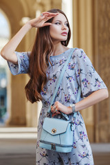 Outdoor portrait of young beautiful fashionable woman posing in street. Model wearing stylish jumpsuit with flower print, wrist watch, a lot of rings, holding light blue bag. Female fashion concept