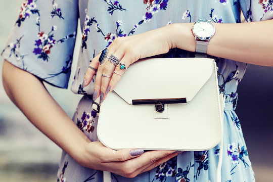 Close up photo of white leather bag in hands of fashionable woman pising in street. Model wearing silvery wrist watch, a lot of rings. Elegant outfit. Female fashion concept