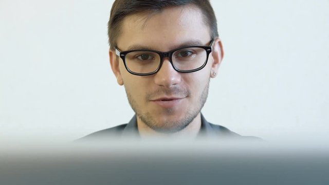 Close-up portrait of a young man wearing glasses sitting in his office in front of a monitor - having a video chat. People stock footage slider shot. 