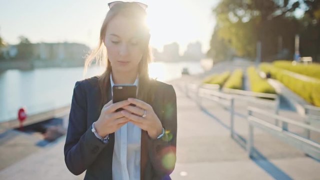 Young Businesswoman Using SmartPhone, Going to Work in the Sunny Morning City. SLOW MOTION. STEADICAM Stabilized Shot. Attractive Professional Business Woman with a cell phone in a city. Lens Flare. 