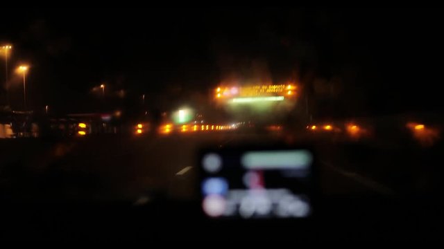 Night road from inside car 4k through defocused GPS navigation device screen. Passing electronic sign board with warning on wet slippery highway in Italy or Switzerland view from drivers perspective