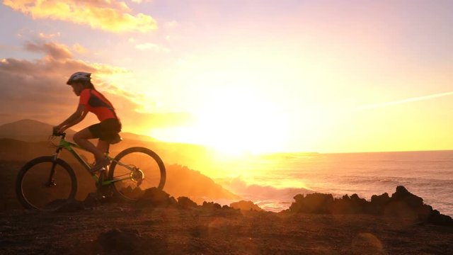 Mountain biking MTB cyclist woman cycling on bike trail at sunset by ocean. Person on bike by sea in sportswear with bicycle enjoying healthy active lifestyle in beautiful nature outside.