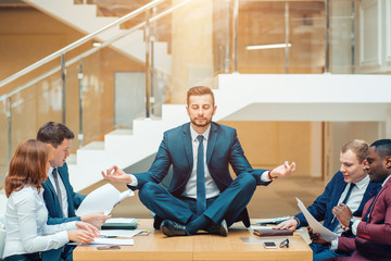 Businessman wearing a suit doing Yoga on the table and other businessmans angry