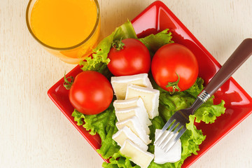 Tomatoes and cheese slices lie on a sheet of fresh salad in a red plate. A glass of orange juice stands side by side on the table.