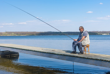 Lonely senior fisherman sitting on wicker stool on a pier with rod and ready to catch fish.