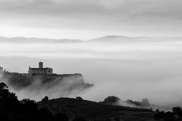 Fototapeta na wymiar Beautiful monochrome view of St.Francis church in Assisi (Umbria, Italy), over a sea of fog at dawn, with hills and trees in the foreground