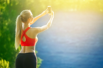 young blonde woman stands on a blue sea background. Sports woman in red t-shirt makes selfie on nature near the sea